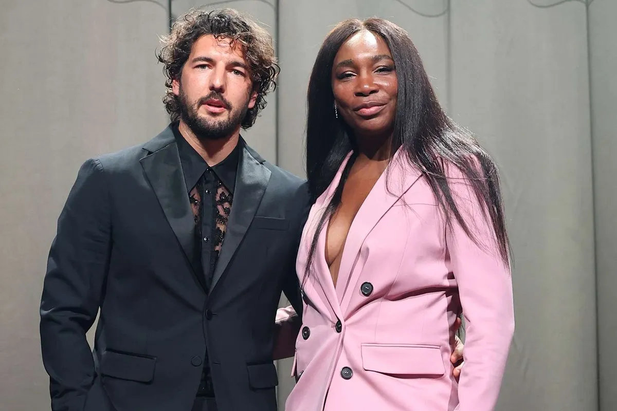 Un uomo in completo nero e una donna in blazer rosa posano insieme su uno sfondo neutro durante un evento di moda