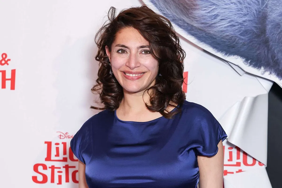 Caterina Murino in primo piano, sorridente, con capelli mossi e abito di raso blu, sullo sfondo di un photowall bianco con loghi in rosso.