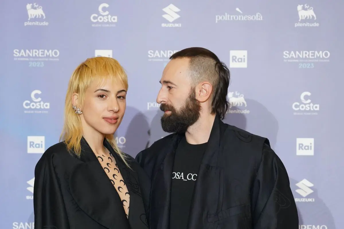 I componenti del duo Coma_Cose (una donna dai capelli biondo platino con ciuffo e un uomo con barba folta e capelli rasati ai lati) in abiti scuri sul red carpet del Festival di Sanremo 2023, con loghi Rai, Sanremo, Costa, Suzuki e Poltronesofà sullo sfondo.