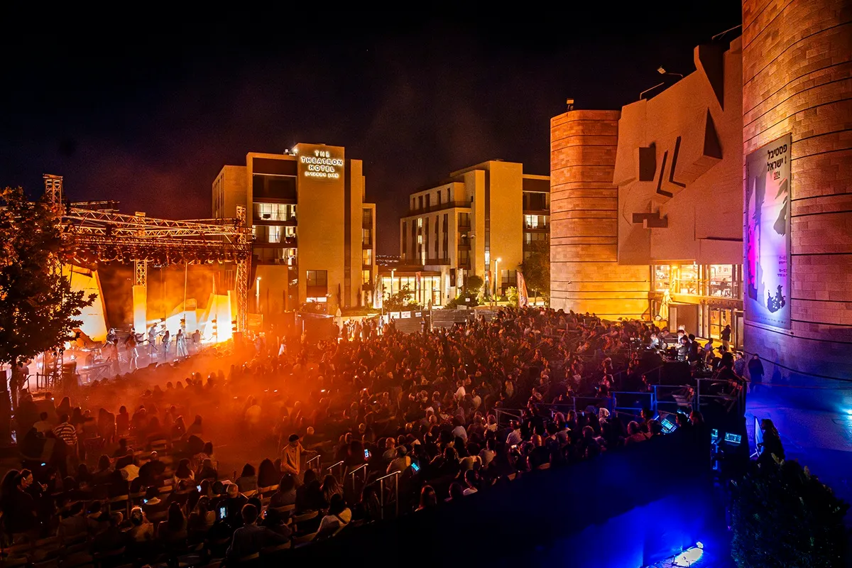 Folla seduta di notte davanti a un palcoscenico illuminato da luci arancioni al Festival d’Israele in Gerusalemme 2025, con edifici moderni sullo sfondo.