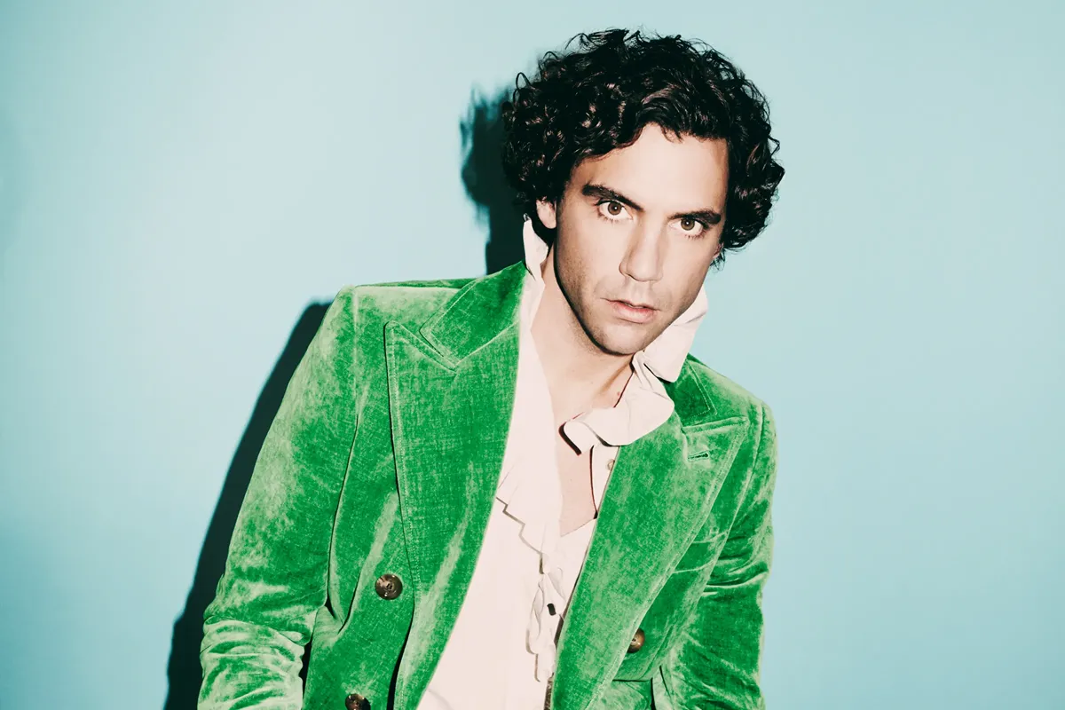 Ritratto di MIKA con giacca di velluto verde su sfondo celeste.