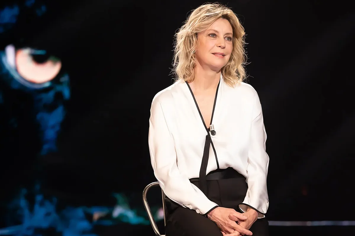 Donna bionda con camicia bianca bordata di nero e microfono a clip seduta su sgabello con sfondo scuro