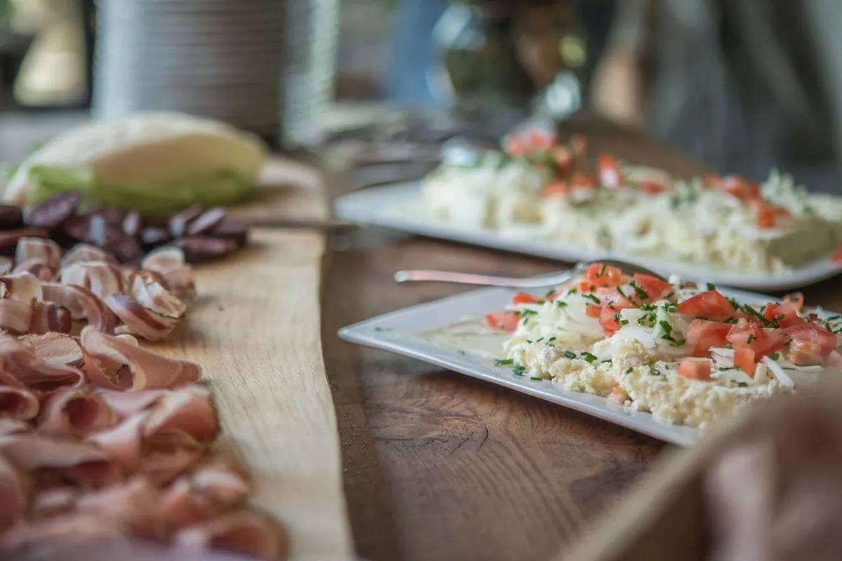 Piatto con Graukäse guarnito con pomodori e erba cipollina accanto a tagliere con speck e salumi tradizionali su tavolo in legno rustico.