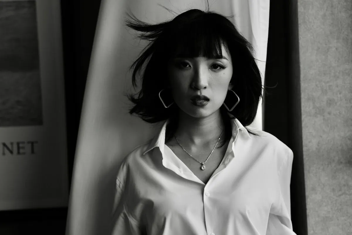 Mulher com cabelo curto escuro e franja, usando uma camicia bianca donna elegante, brincos grandes e colar, em foto preto e branco com fundo escuro.