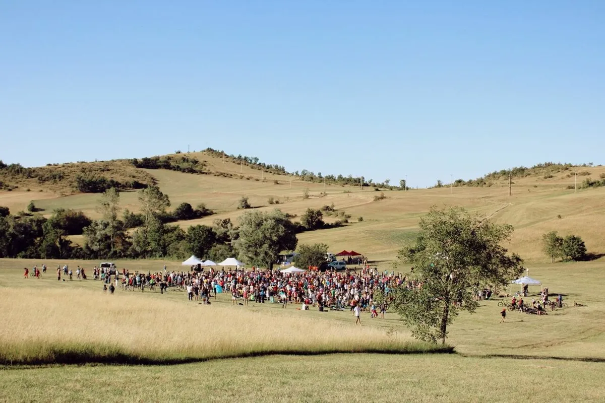 Gruppo di centinaia di persone riunite su un prato verde tra dolci colline delle Marche durante il festival IncantoMarche, con gazebo e biciclette sotto un cielo sereno
