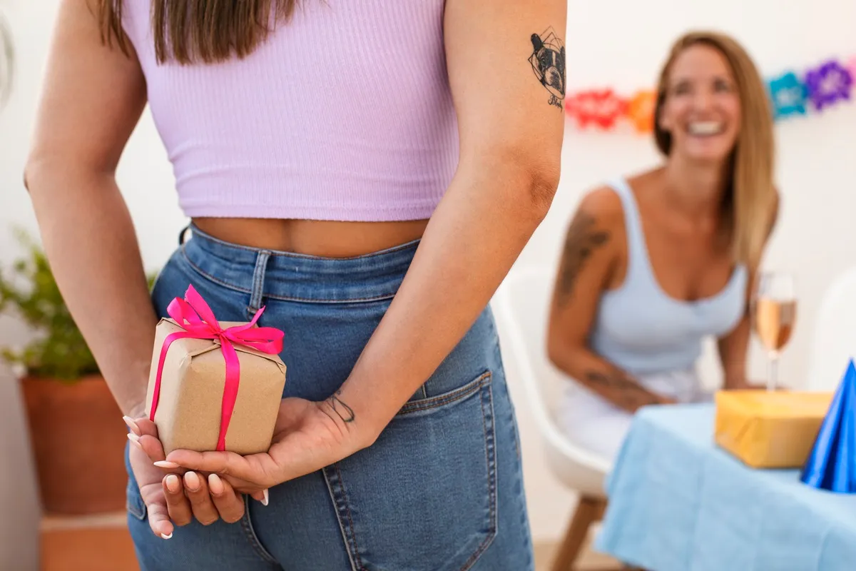 Donna con canotta lilla tiene un pacchetto regalo avvolto in carta marrone e nastro rosa dietro la schiena, mentre un’amica sorridente è seduta a un tavolo decorato per la festa.