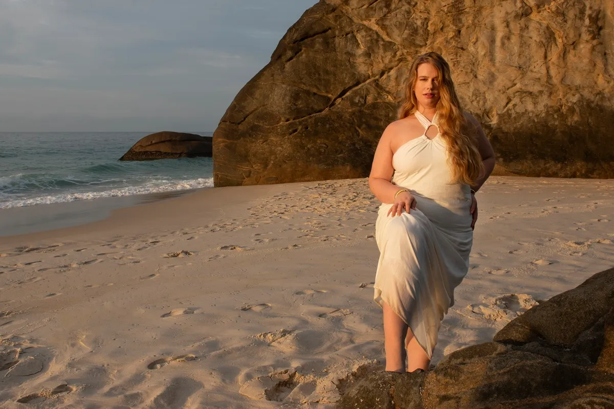 Donna curvy in abito da cerimonia bianco con scollo ad anello, posa su una spiaggia sabbiosa davanti a grandi rocce.