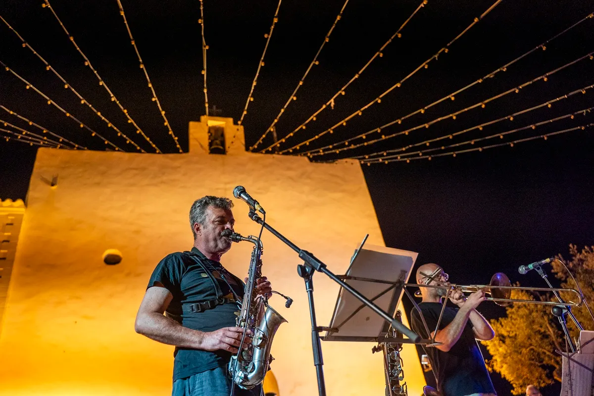Saxofonista in primo piano suona di notte davanti a un edificio storico illuminato, con file di lucine sospese che convergono al centro del cielo scuro.