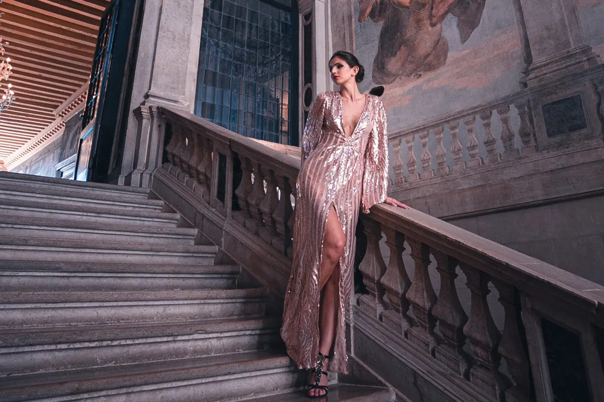 Donna in abito lungo rosa con paillettes appoggiata a una maestosa scalinata in un interno di palazzo storico.