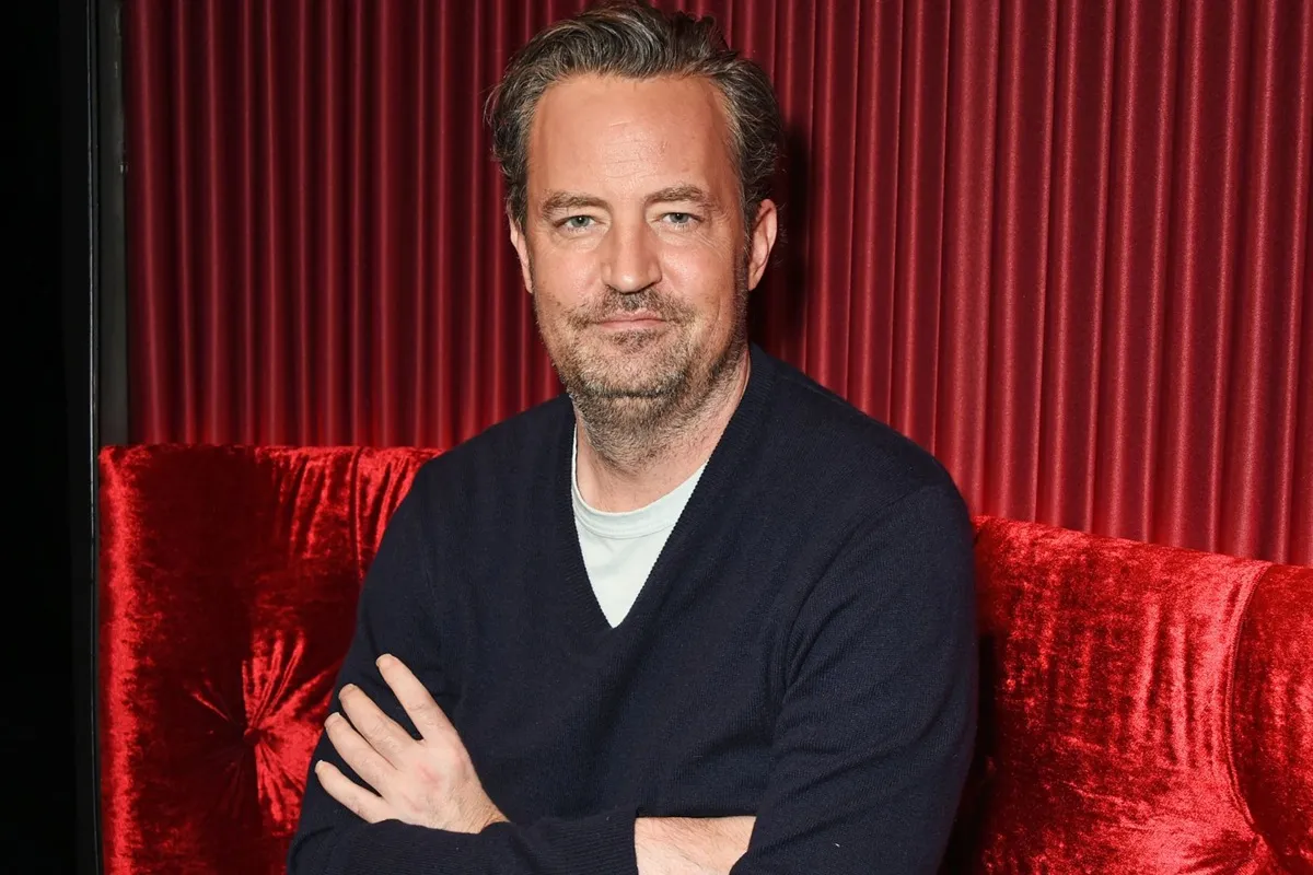 Primo piano di Matthew Perry seduto su un divano di velluto rosso, indossa un maglione scuro e guarda l’obiettivo con espressione pensierosa.