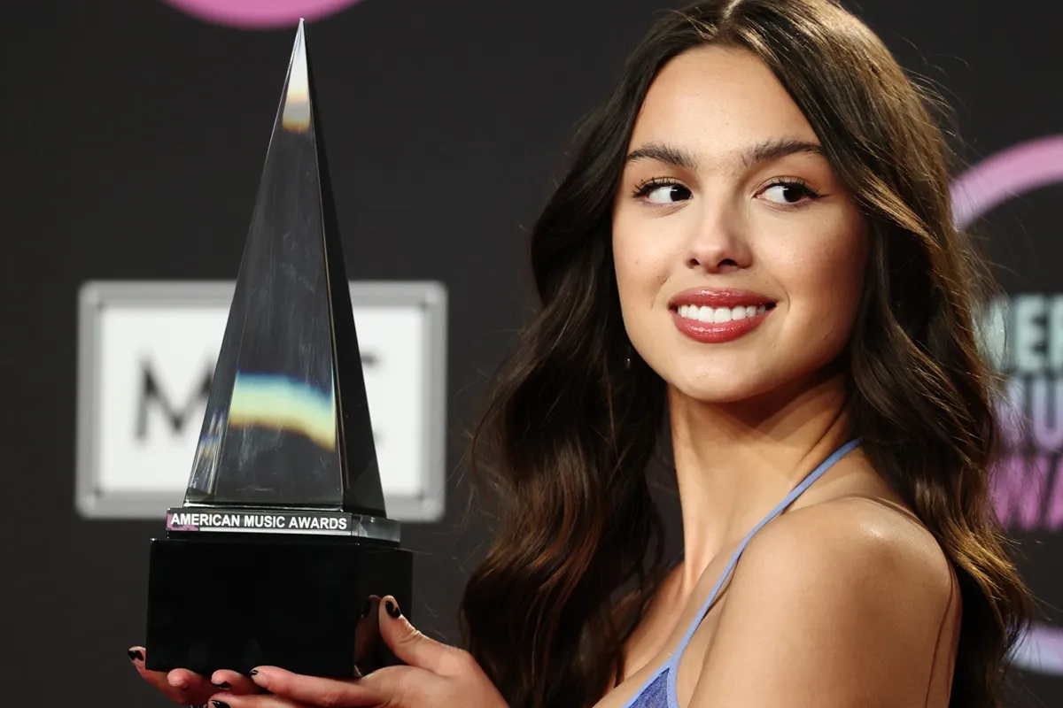 Olivia Rodrigo sorride tenendo un trofeo triangolare degli American Music Awards sul palco, sfondo scuro con dettagli rosa.