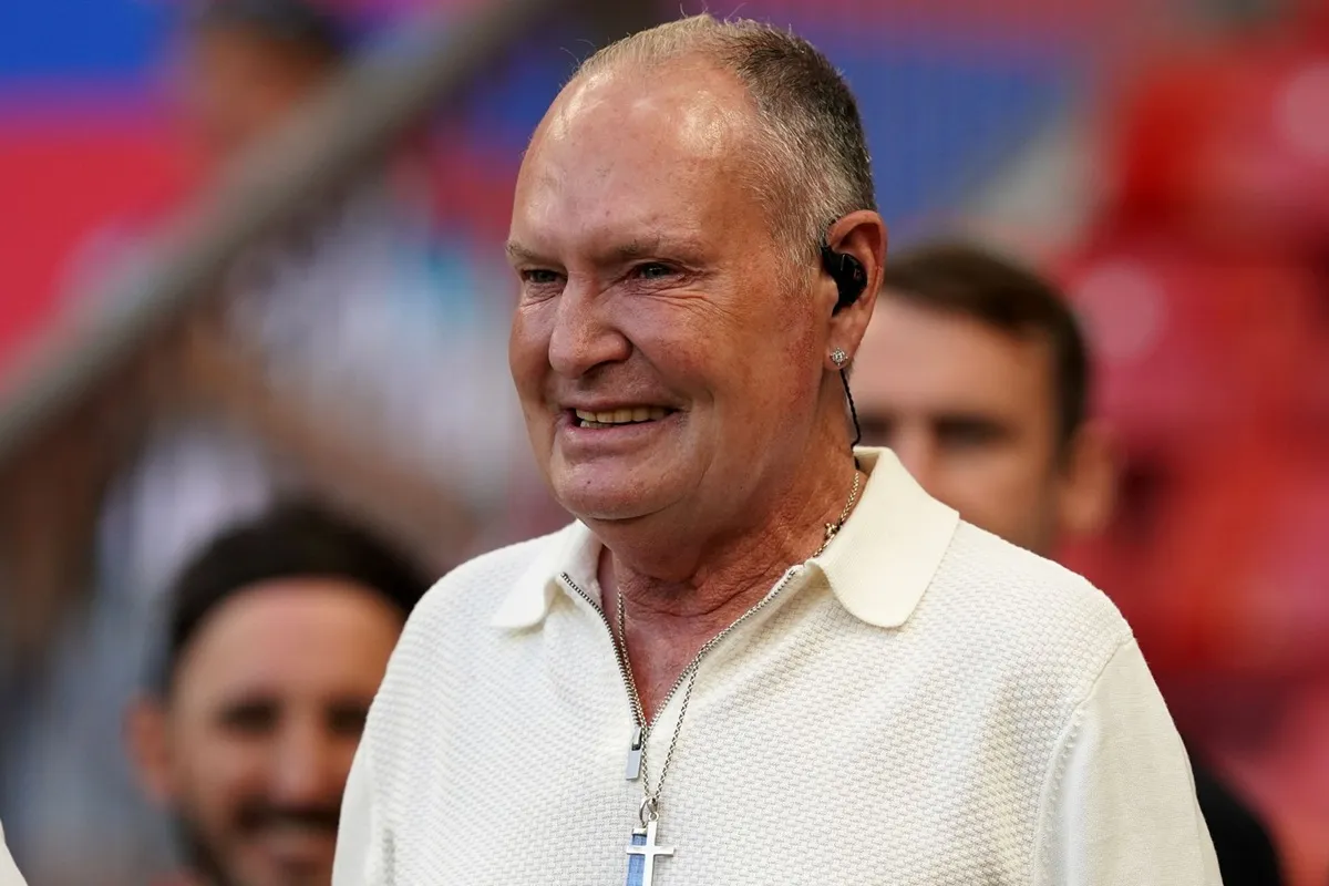 Primo piano di un uomo di mezza età che sorride, indossa un polo bianco, un auricolare nero e una collana con un ciondolo a forma di croce, sullo sfondo di uno stadio affollato.