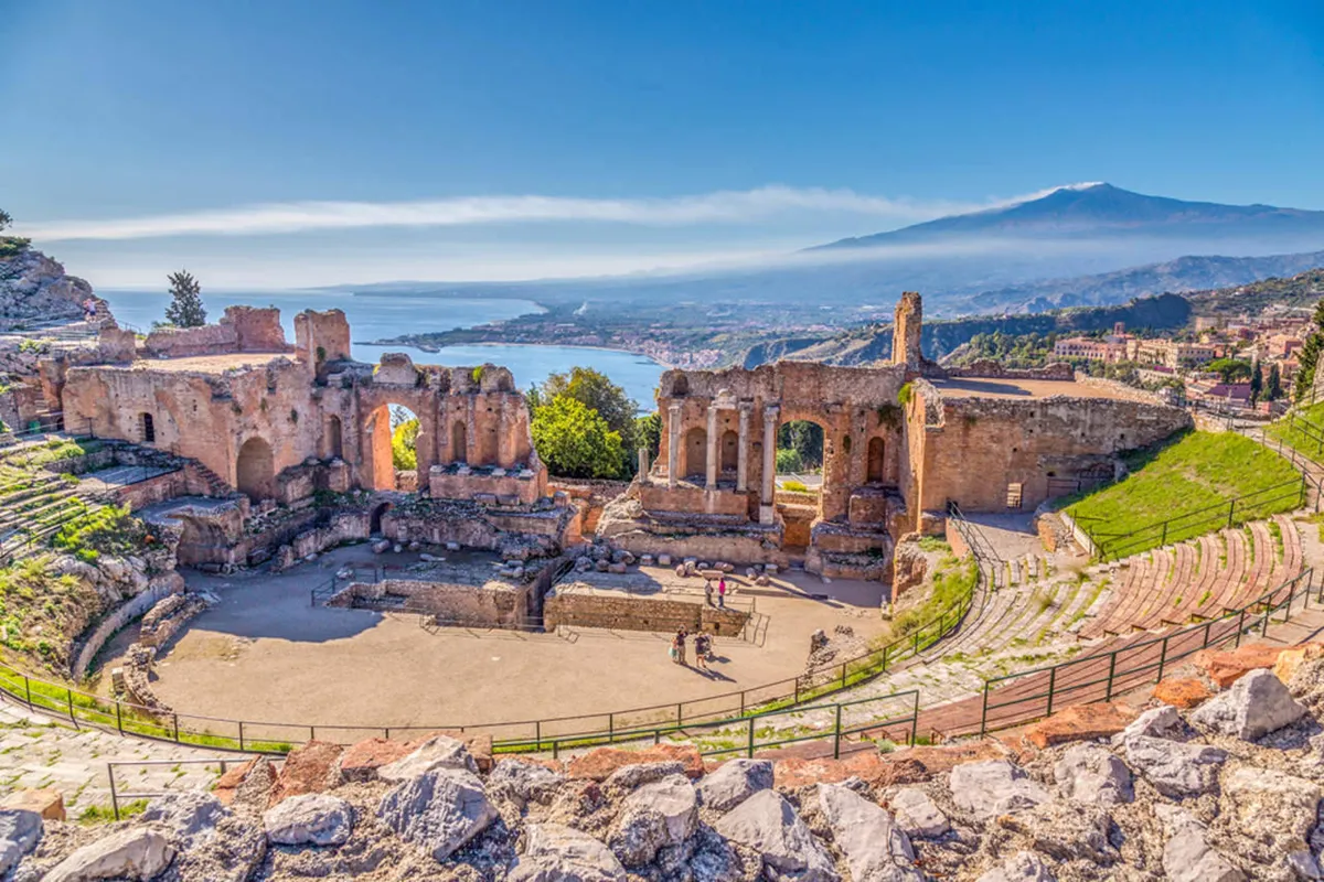 Teatro antico all’aperto con gradinate in pietra, rovine archeologiche, mare sullo sfondo e il vulcano Etna all’orizzonte