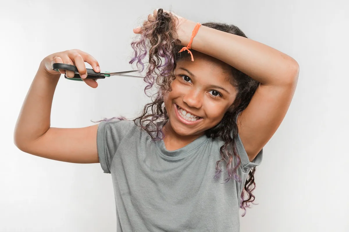 Ragazza sorridente con capelli ricci che si taglia la chioma con delle forbici