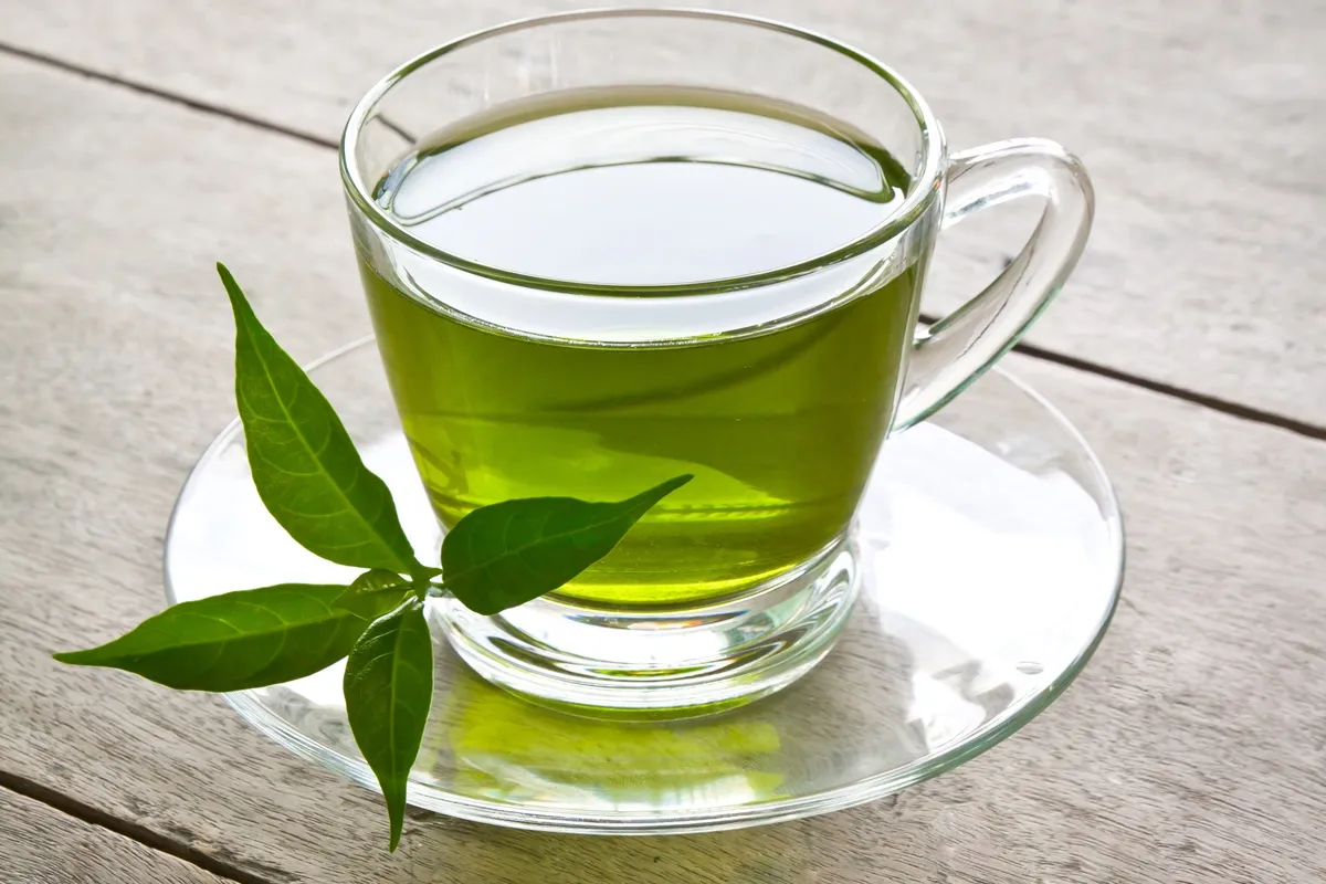 Tazza di tè verde in vetro trasparente con foglie di tè fresche poggiata su un tavolo di legno chiaro