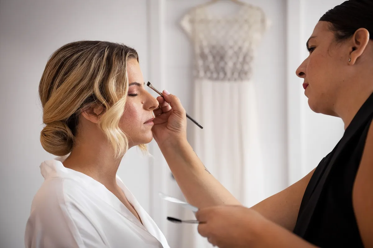 Make-up da sposa in corso: un’artista del trucco applica ombretto rosa chiaro alla sposa, con un abito bianco appeso sullo sfondo.