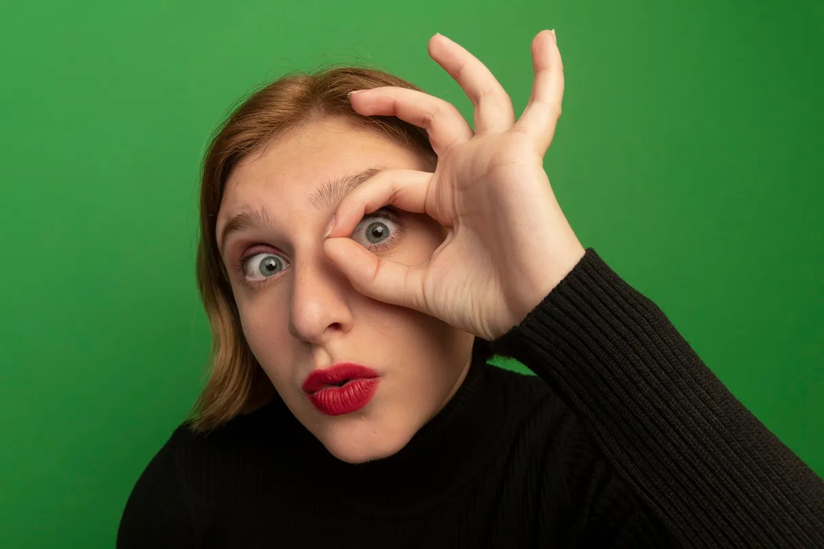 Primo piano di una donna con occhi verdi e rossetto rosso, sfondo verde brillante, che forma un cerchio con le dita attorno all’occhio destro