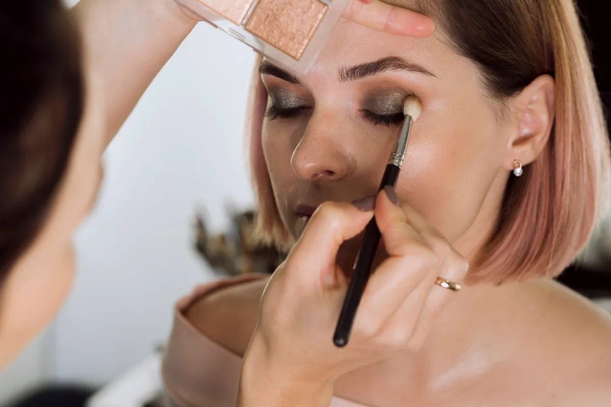 Make-up artist applica ombretto scuro su palpebra di una donna con pennello, vicino a una palette di tonalità neutre