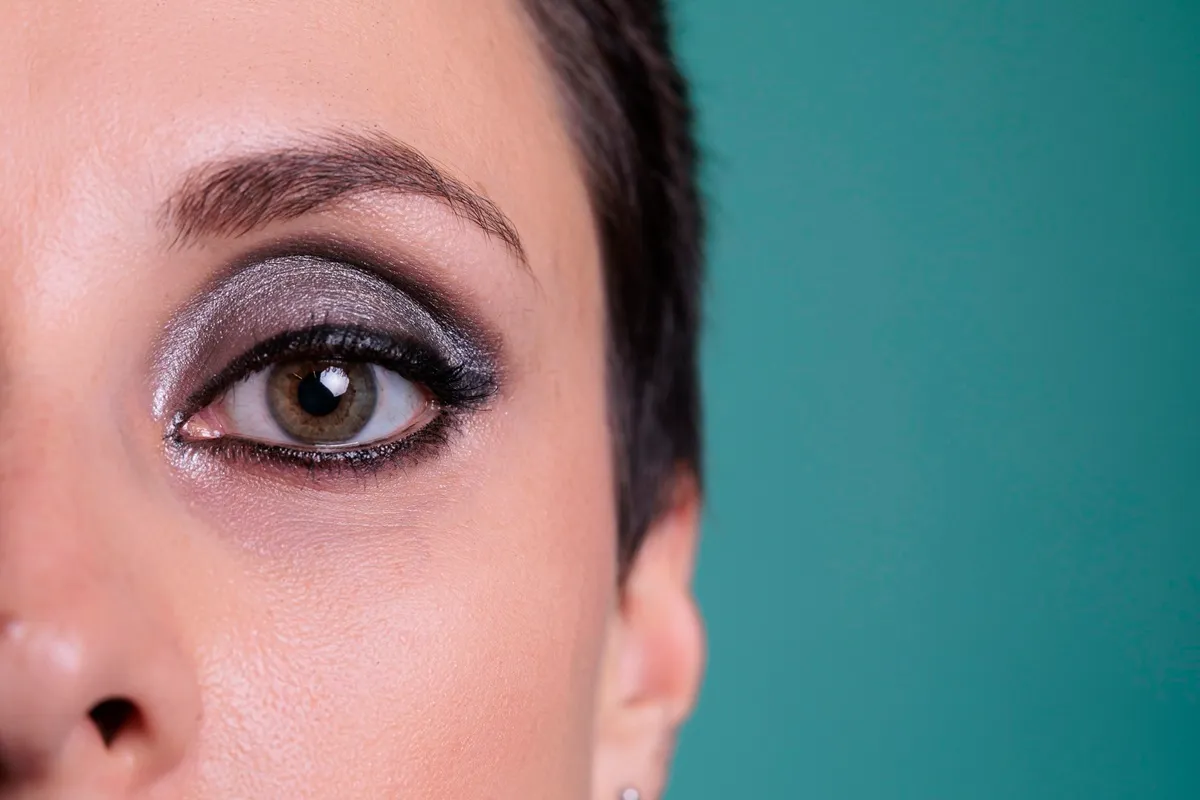 Olho feminino com maquiagem smokey eye prateada e delineado preto, sobrancelha definida e fundo verde