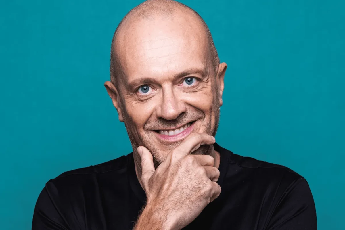 Primo piano di un uomo calvo con occhi azzurri e sorriso aperto, con una mano appoggiata sul mento e sfondo turchese uniforme.