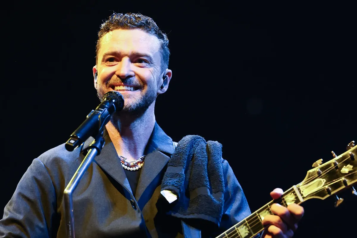 Uomo sorridente al microfono con chitarra elettrica in mano, illuminato sul palco contro sfondo nero.