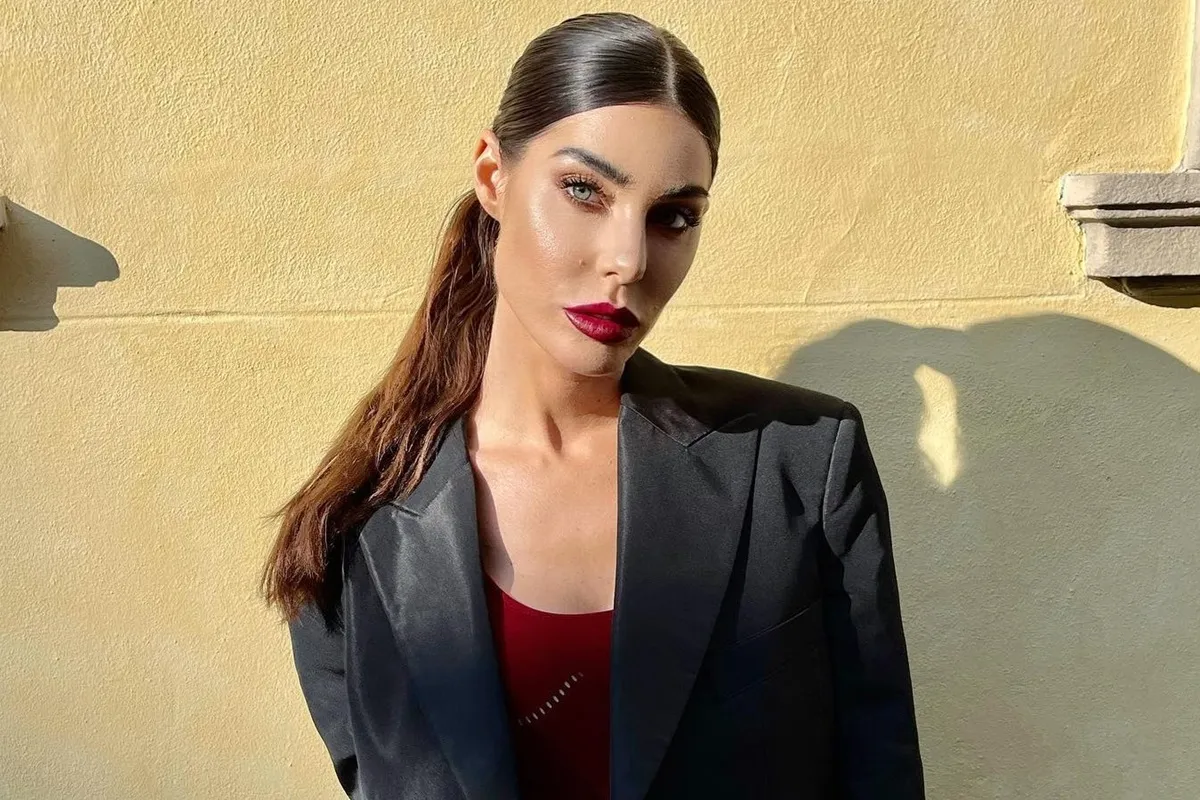 Bianca Atzei posa con trucco elegante e rossetto rosso intenso, indossando giacca nera e top rosso, davanti a una parete color crema illuminata dal sole.
