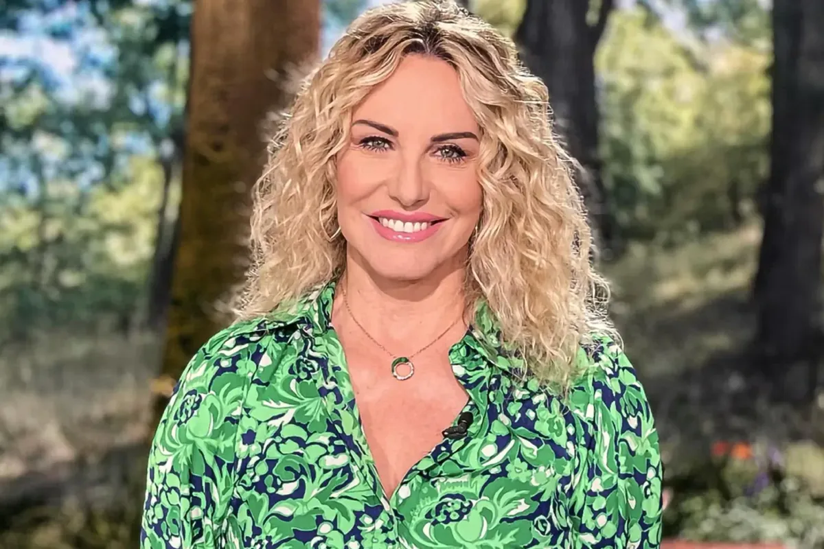 Primo piano di donna bionda con capelli ricci, camicia verde con motivo floreale e collana sottile, ritratta all’aperto con alberi sfocati sullo sfondo