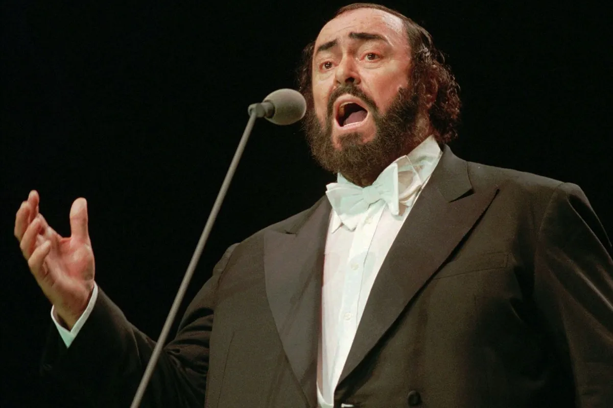 Luciano Pavarotti in giacca da sera nera e papillon bianco, canta appassionato di fronte a un microfono sul palco.