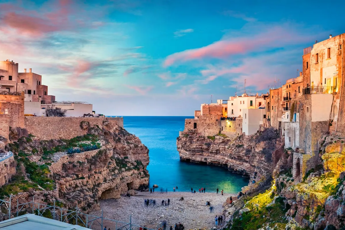 Scogliera calcarea che delimita una piccola spiaggia ghiaiosa con acqua turchese, edifici in pietra chiara ai lati e cielo al tramonto dai toni rosa.