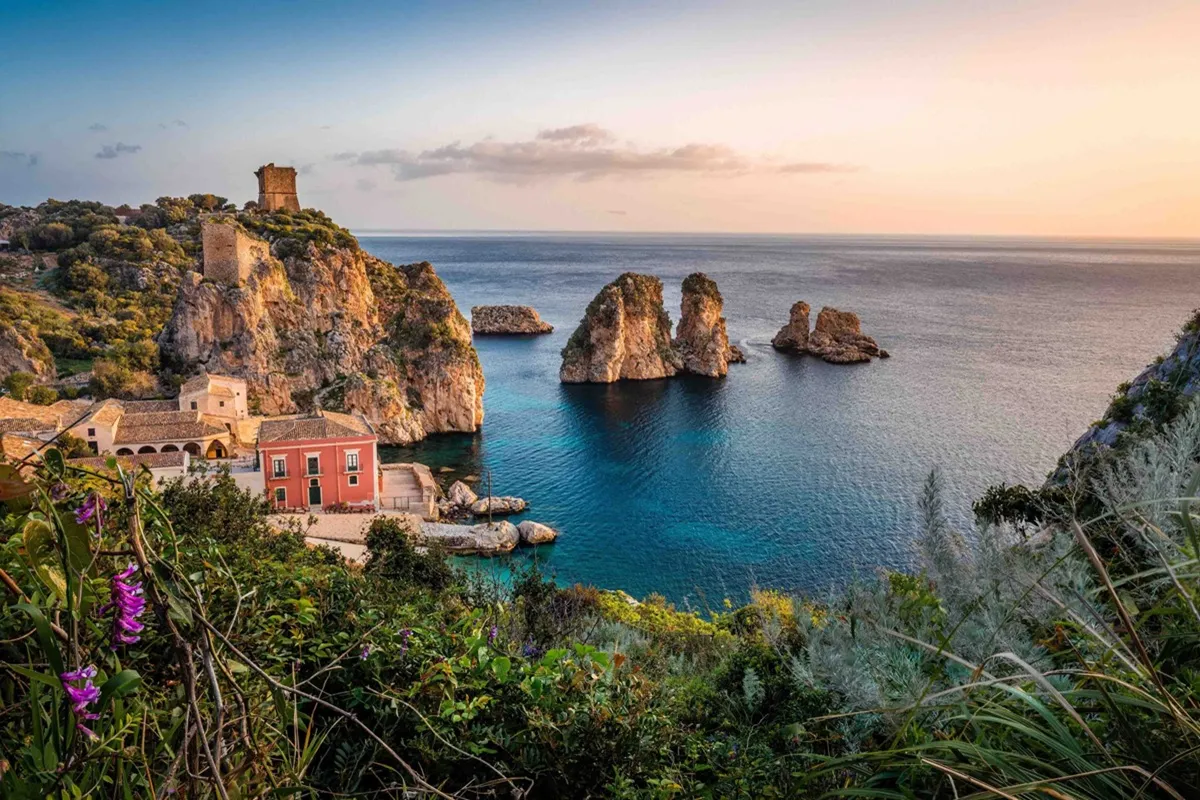 Veduta dei faraglioni di Scopello al tramonto con il borgo storico costiero e il mare turchese
