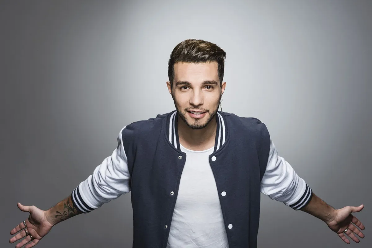 Primo piano di un giovane uomo sorridente in giacca varsity blu e bianca, con le braccia aperte su sfondo grigio
