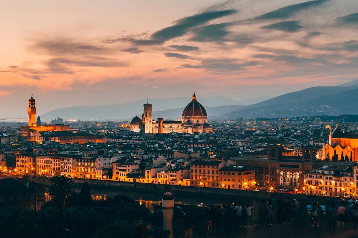 Panorama di Firenze al crepuscolo: la Cattedrale di Santa Maria del Fiore con cupola, il Campanile di Giotto e Palazzo Vecchio illuminati, vista sul Lungarno e sulle colline sullo sfondo
