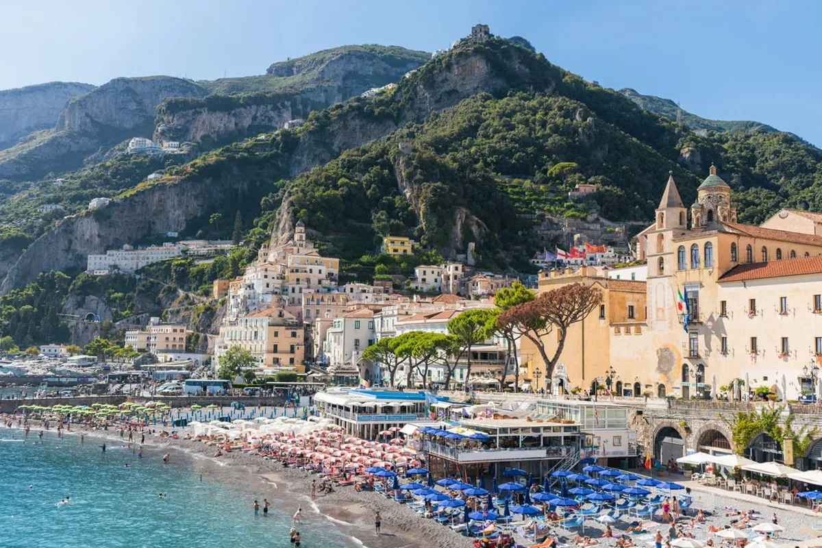 Veduta panoramica della spiaggia di Amalfi con ombrelloni colorati, mare turchese e case arroccate sulle colline della Costiera Amalfitana.