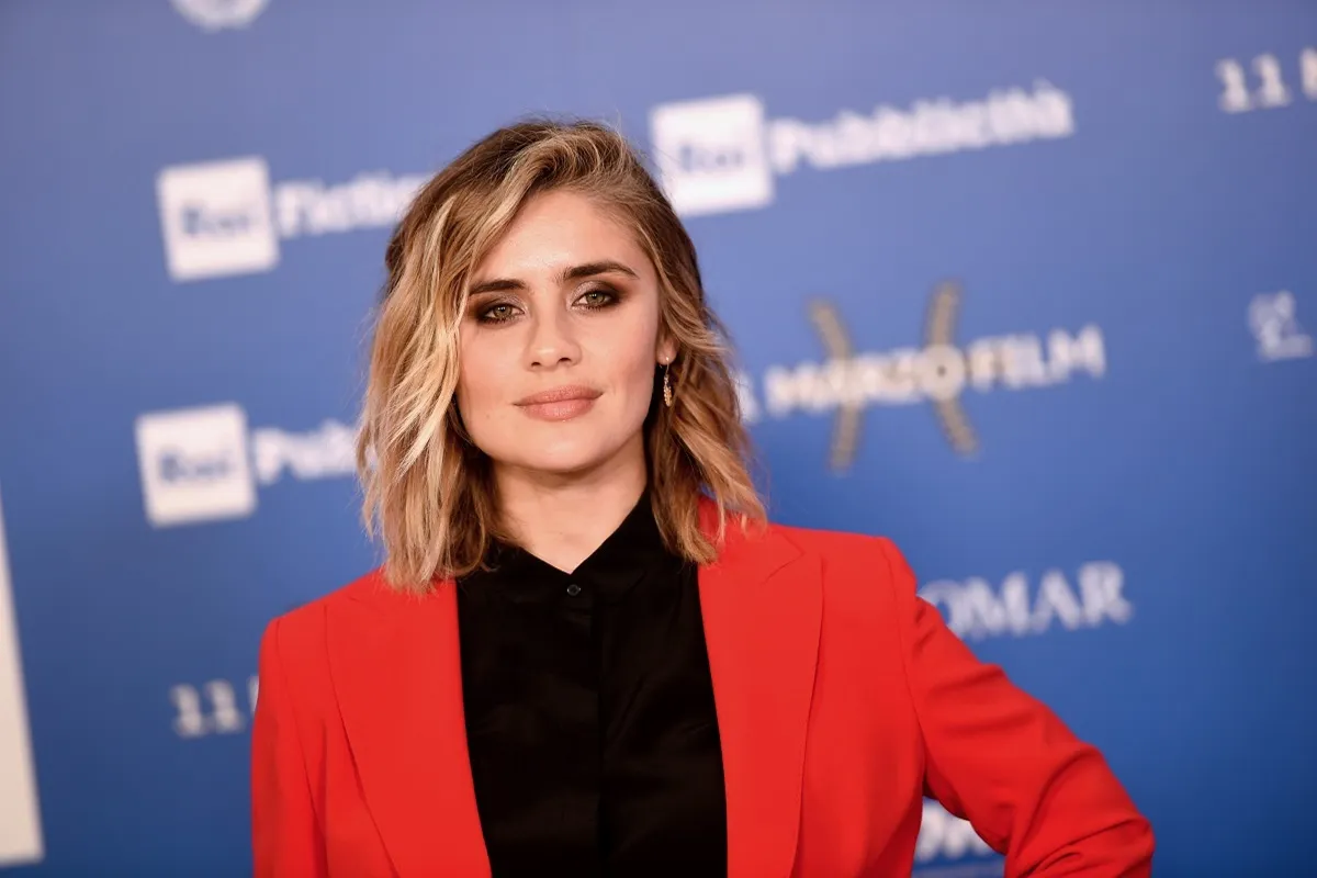 Attrice italiana posa sul red carpet indossando una giacca rossa elegante davanti a un pannello blu con loghi di sponsor durante un festival del cinema.