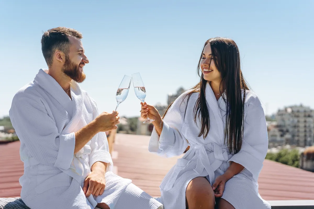 Coppia sorridente in accappatoi bianchi che brinda con calici di prosecco su una terrazza soleggiata durante un weekend romantico in spa.