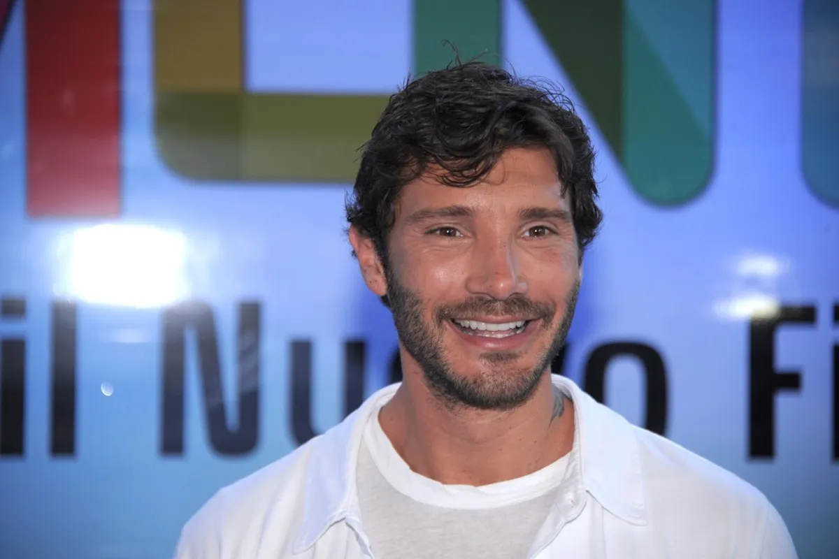 Stefano De Martino sorride di fronte alle telecamere, indossando una camicia bianca sopra una t-shirt, durante un evento pubblico in Italia.