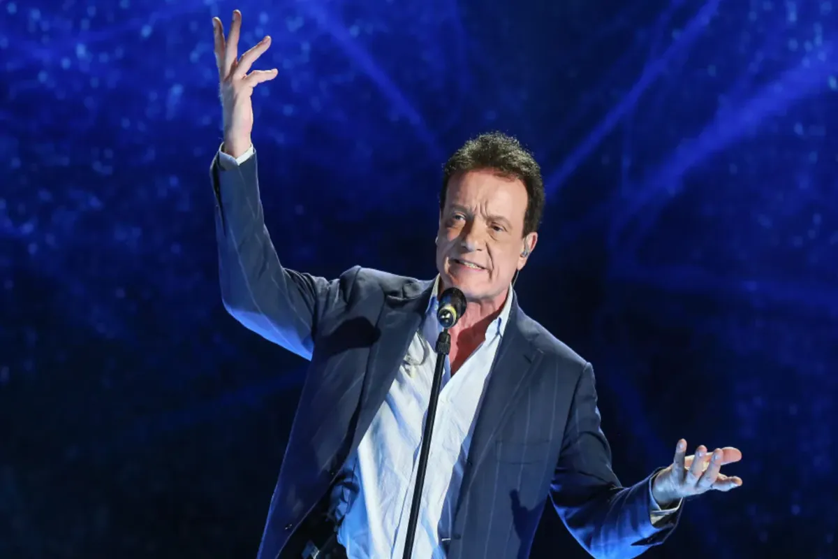 Massimo Ranieri in abito elegante canta al microfono con le braccia aperte davanti a uno sfondo blu luminoso durante un concerto dal vivo