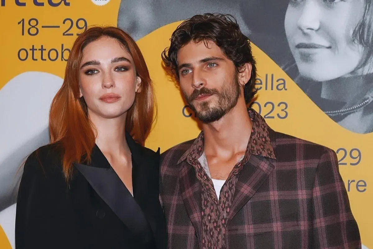 Coppia vestita elegantemente con tailleur nero e giacca a quadri in posa davanti a poster giallo con date di festival.