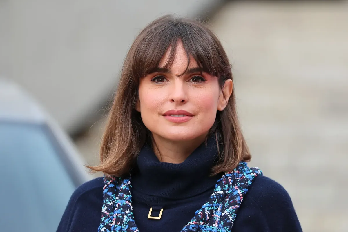Verónica Echegui sorride con un look elegante, indossando un maglione blu scuro con dettagli colorati e capelli castani con frangia, in uno scatto a mezzo busto all’aperto.