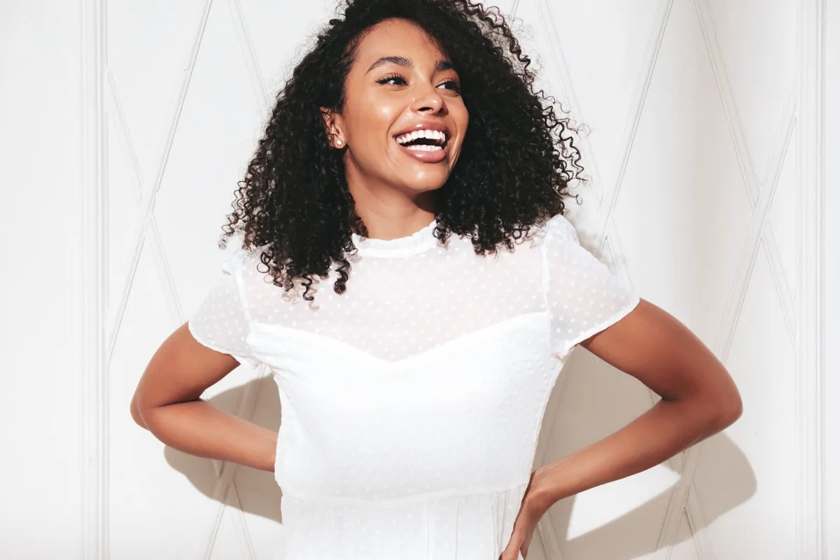 Donna sorridente con capelli ricci naturali, indossa un abito bianco trasparente a pois sopra il corpetto, posa luminosa e solare.