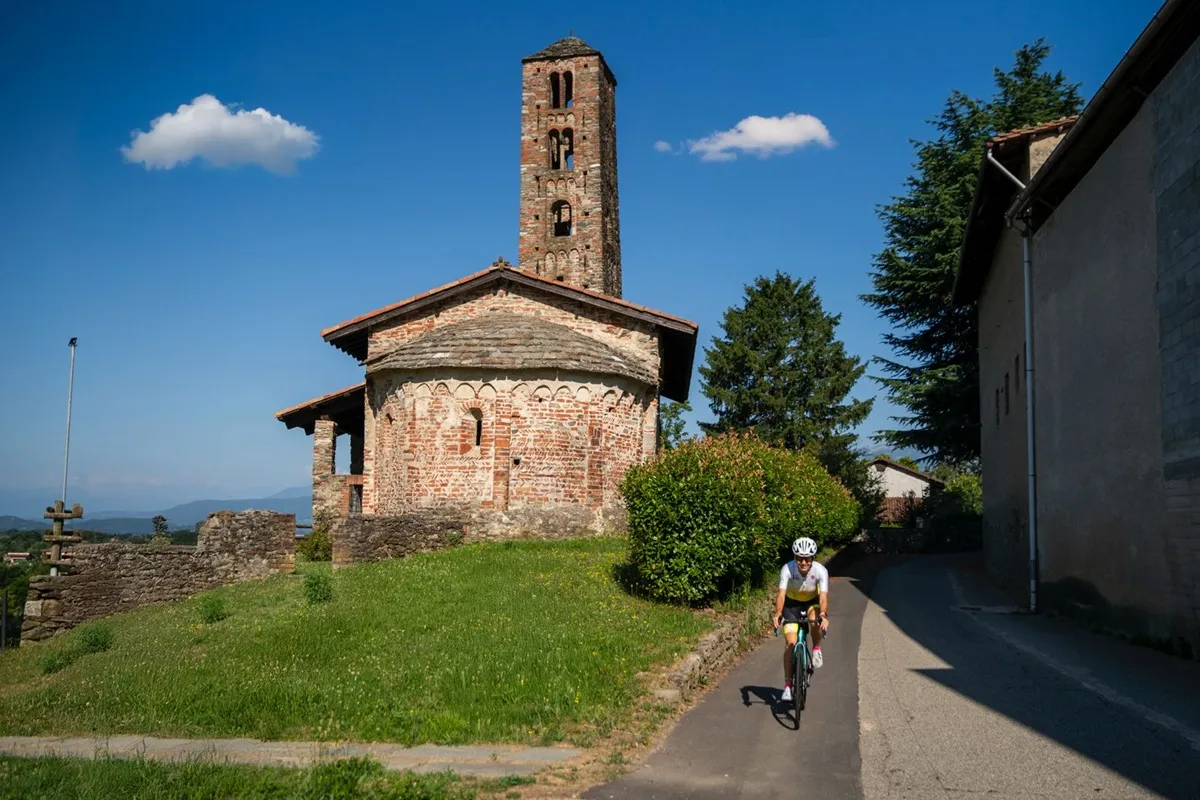 Ciclista durante il percorso del 100x100 Donne a Ivrea passa davanti a una chiesa romanica in mattoni nel Canavese sotto un cielo azzurro.