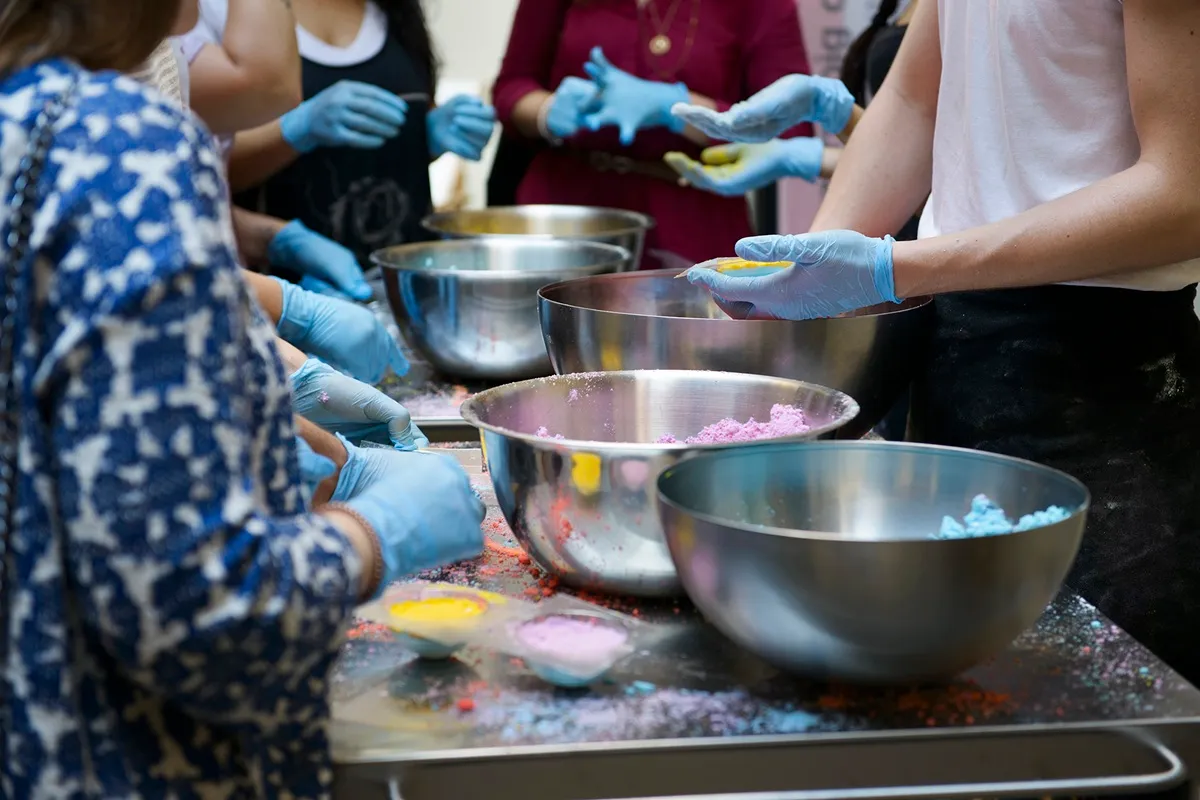 Persone con guanti blu partecipano a un laboratorio creativo, mescolando polveri colorate in grandi ciotole di acciaio inox.