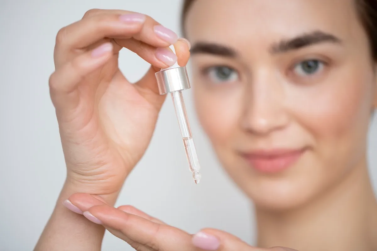 Primo piano di una giovane donna con pelle luminosa che tiene un contagocce con siero trasparente di acido ialuronico, pronta ad applicarlo sulla mano per la cura del viso.