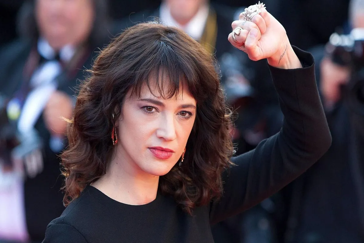 Asia Argento con i capelli castani mossi e frangia, vestita di nero, alza il pugno con un’espressione decisa durante un evento pubblico.