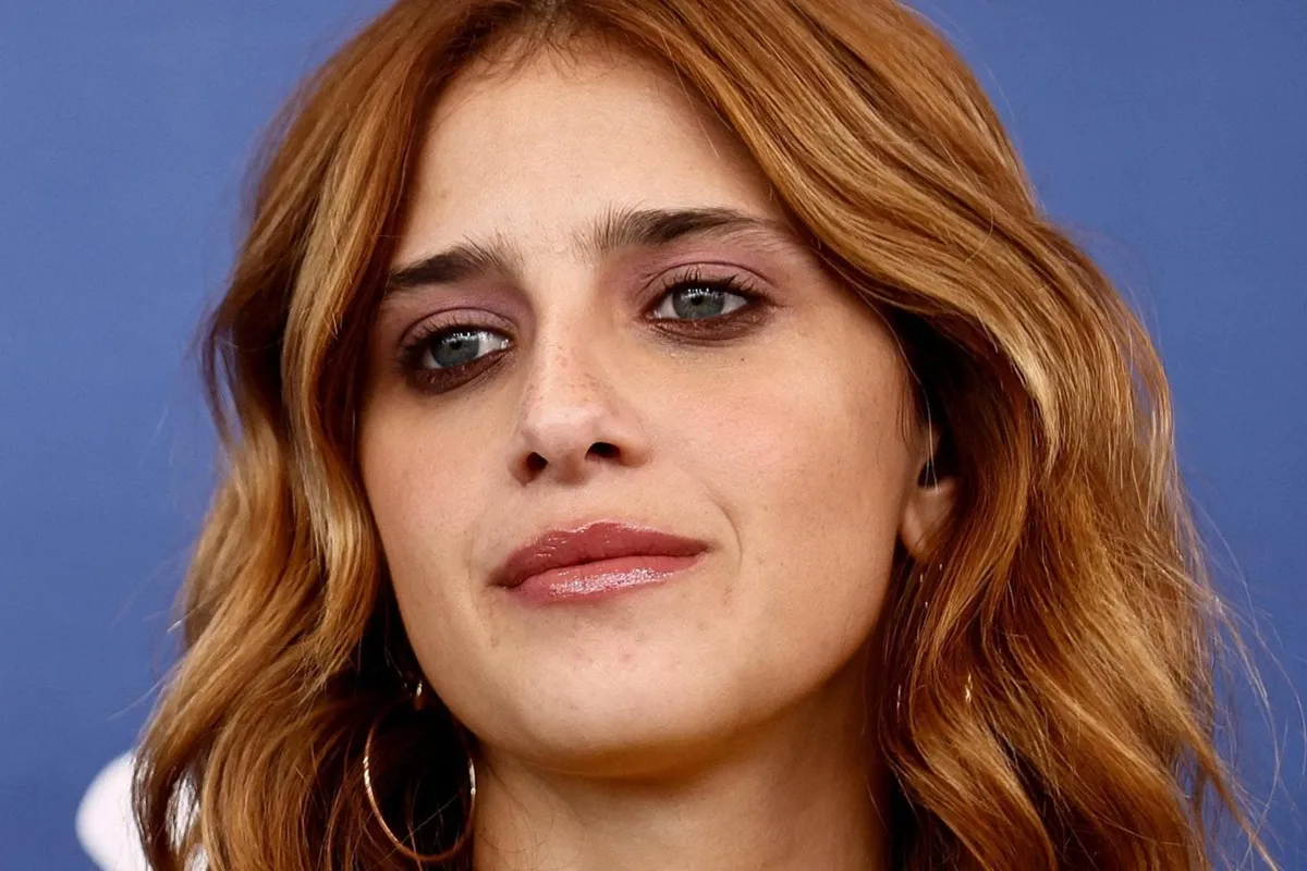 Primo piano di Benedetta Porcaroli con capelli castano-ramati ondulati, trucco naturale e sguardo profondo su sfondo blu.