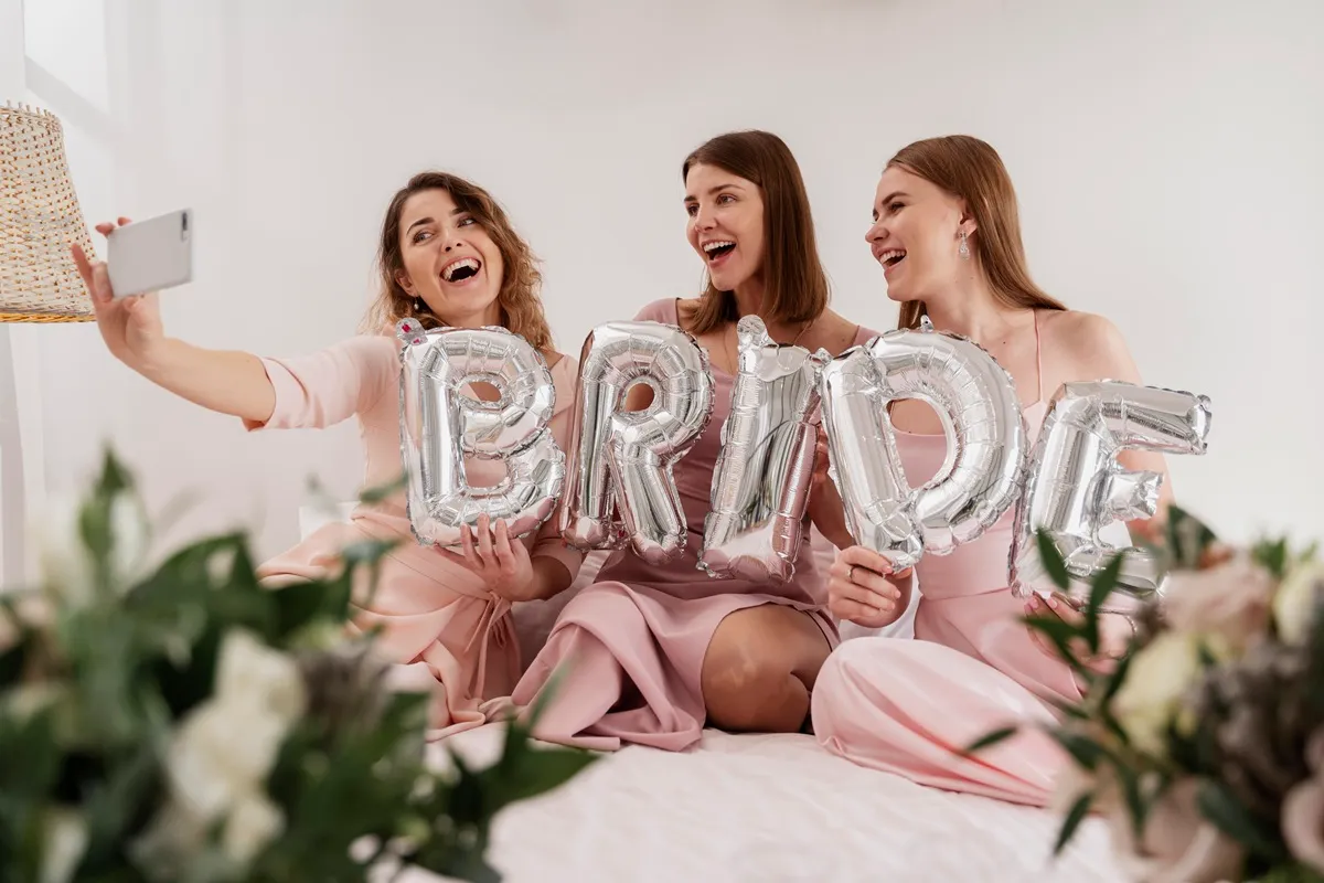 Tre donne sorridenti in abiti rosa scattano un selfie durante un addio al nubilato, tenendo palloncini argentati con la scritta Bride.