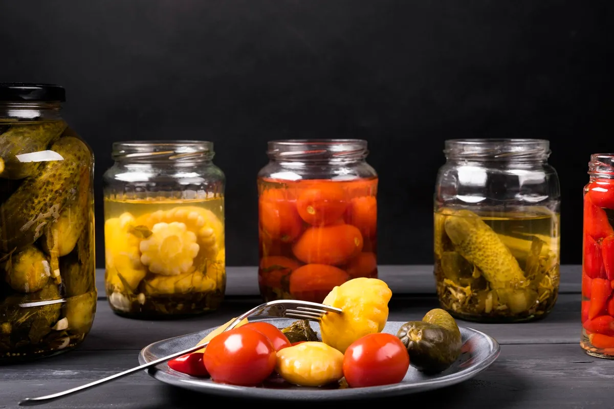 Barattoli di vetro con verdure fermentate e sott’aceto, tra cui cetrioli, pomodori e cavolfiori, con un piatto di assaggi in primo piano.