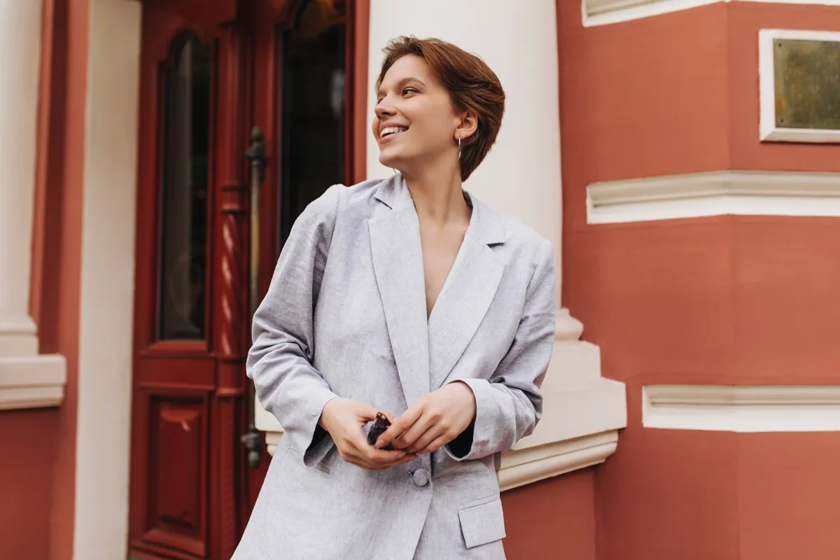 Donna sorridente con capelli corti e blazer oversize grigio chiaro, posa davanti a un edificio storico color terracotta e bianco.