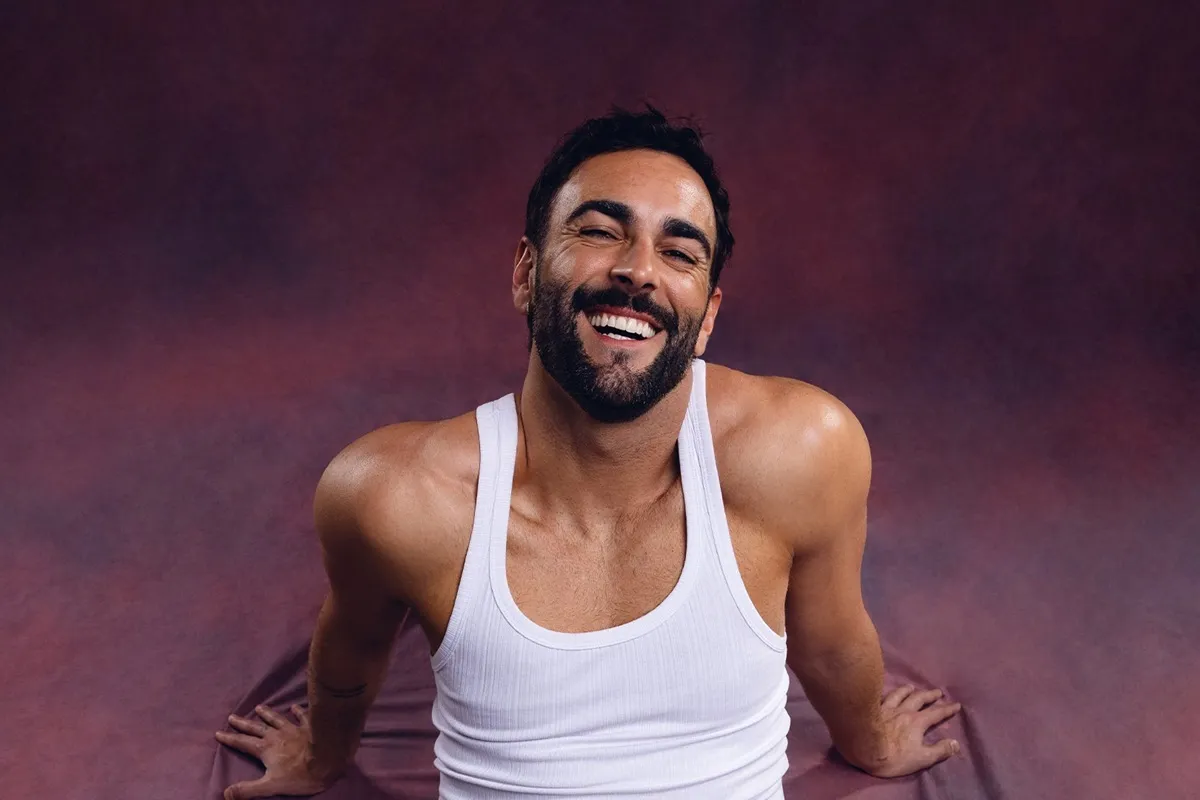 Uomo con barba e capelli scuri, indossa una canotta bianca e sorride mentre posa su sfondo viola sfumato.