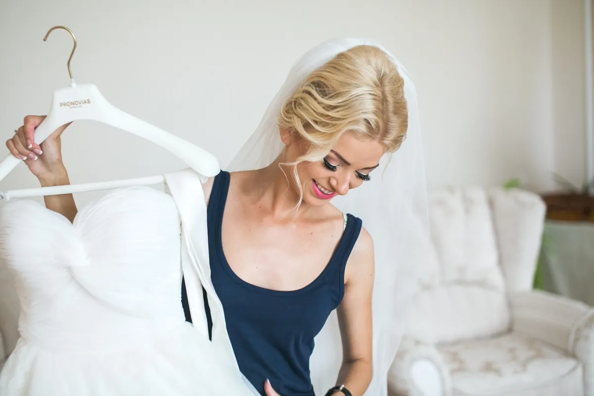 Giovane sposa bionda con velo sorride mentre tiene in mano il suo abito da sposa bianco su una gruccia.